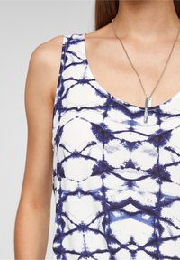 Vit linne med ett blått abstrakt mönster, med rund halsringning och design med snörband. Silverhalsband med en rektangulär berlock.