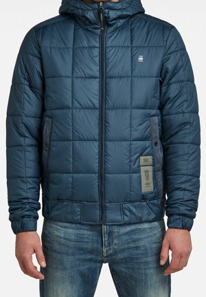 Mand iført en blå quiltet hættetrøje med lynlås foran og sidelommer, kombineret med blå jeans mod en hvid baggrund.