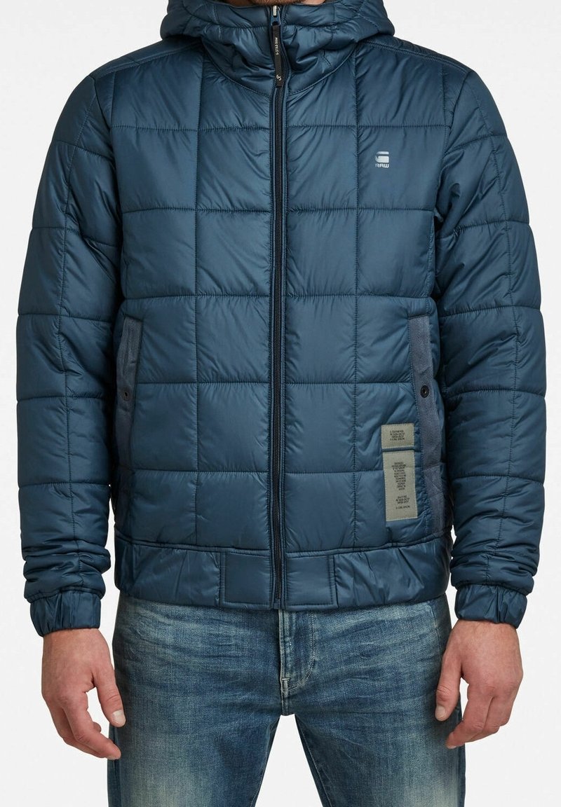 Homme portant une veste matelassée bleue à capuche avec fermeture éclair devant et poches latérales, associée à un jean bleu sur fond blanc.