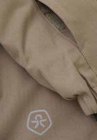 Hellbraunes, in Fischgrätmuster gewebtes Material mit strukturierter Oberfläche. Über ein Logo in einer helleren Nuance in der unteren linken Ecke und eine offene Tasche.