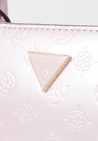 Borsa a tracolla rosa goffrata con logo triangolare Guess, caratterizzata da motivi delicati e una texture liscia, accentuata da dettagli in oro.