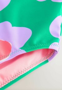 Fürdőruha anyag nagy zöld háttérrel, lila és rózsaszín virágmintákkal. Jellemzője a sima textúra, rózsaszín bélés és varrott szélek.