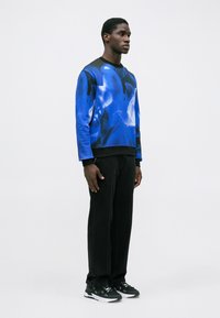 Hombre de pie vestido con un sudadera azul y negra de patrón abstracto, pantalones anchos negros y zapatillas en blanco y negro.