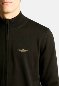 Maglione nero con cerniera frontale e collo alto a coste, caratterizzato da un piccolo emblema dorato con ali ricamato sul petto. Realizzato in tessuto a maglia.