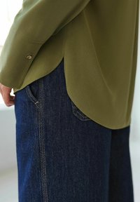 Blouse vert olive en tissu léger, avec un ourlet arrondi et un accent doré sur les poignets, associée à un jean en denim bleu foncé avec des poches visibles.