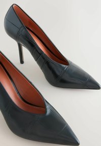 Fekete bőr, hegyes orrú magassarkú cipők, varrott mintával és barna belső béléssel, világos háttér előtt.