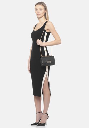 Kobieta w czarnej, bezrękawnikowej sukience midi z białym paskiem po boku zapinanym na guziki, trzymająca czarną torebkę z złotym łańcuszkiem, nosząca czarne szpilki.