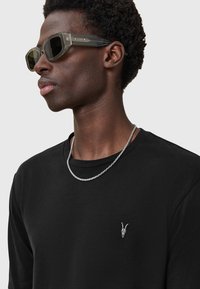 Schwarzes Langarmshirt mit einer kleinen weißen Ziegenschädel-Stickerei, silberner Kettenhalskette und grauen Sonnenbrille mit geometrischem Design.