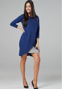 Long-sleeve, navy blue dress made of soft fabric, featuring a draped design and a gray skirt panel. Worn with black high-heeled shoes.