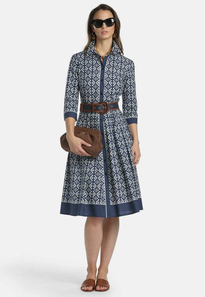 Frau trägt ein blau gemustertes knielanges Kleid mit 3/4-Ärmeln, braunem Gürtel und Clutch, braunen Sandalen, schwarze Sonnenbrille und goldene Ohrringe.