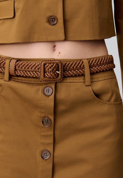 Giacca cropped marrone con grandi bottoni circolari, abbinata a una gonna aderente con una cintura intrecciata marrone e cuciture a contrasto.