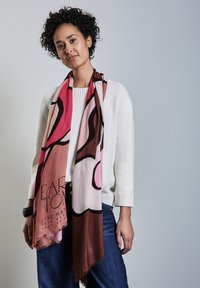 Sciarpa multicolore nei toni di rosa, marrone e beige, con design astratti e bordi frangiati, indossata sopra un maglione bianco con polsini a costine.