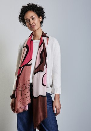Multikleurige sjaal in roze, bruin en beige met abstracte ontwerpen en franjes, gedragen over een witte trui met geribde manchetten.