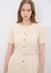 Beige texturované šaty s krátkými rukávy, zlatými knoflíky a odpovídajícím páskem. Mají kulatý výstřih a strukturovanou siluetu.