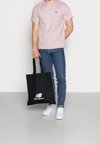 Muž v růžovém tričku Levi's, modrých džínech a bílých teniskách držící černou tašku New Balance na jednobarevném pozadí.