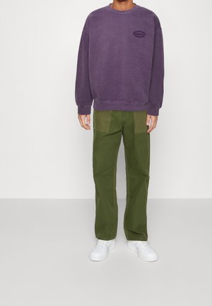Sudadera morada con cuello redondo y logo, combinada con pantalones de pierna ancha en color verde oliva con parches en contraste. Zapatillas blancas completan el look.