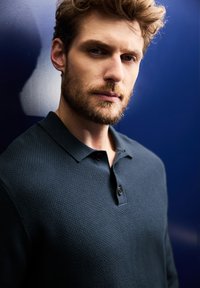 Dunkelblaues, strukturiertes Poloshirt mit Kragen und drei Knöpfen, getragen von einem Model vor einem einfarbigen Marine-Hintergrund.