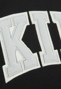 Schwarzes Kleidungsstück mit großen, gestickten Buchstaben in Weiß und einer strukturierten Oberfläche, die hervorstechende Nähte und scharfe Kanten betont.