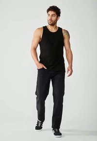 Uomo con capelli ricci che indossa una maglietta nera senza maniche, pantaloni neri e sneakers nere, in piedi con una mano in tasca, che guarda a destra.