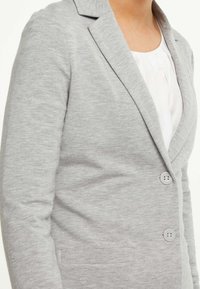 Blazer gris ajusté avec une texture subtile, revers à narines, deux boutons sur le devant et poches latérales, porté sur un chemisier blanc.