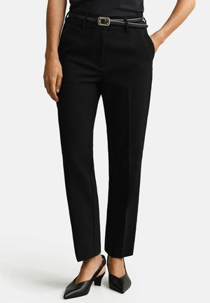 Femme portant un pantalon noir sur mesure, une ceinture noire avec boucle dorée, et des escarpins noirs à brides arrière, debout avec les mains dans les poches.