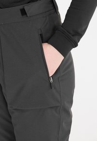 Zwarte skibroek met een gladde textuur, voorzien van een zijzak met rits, versterkte naden en een klittenbandversteller bij de taille.