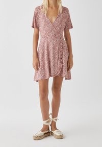 Vestido envolvente rojo con flores y mangas cortas, que presenta un escote en V y un dobladillo con volantes. Se lleva con sandalias de esparto de color claro.