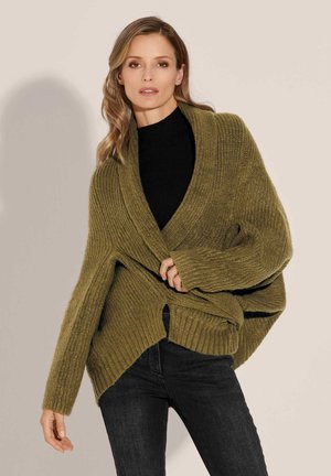 Olivgrüner Strickcardigan mit strukturiertem Rippmuster, oversized Passform, V-Ausschnitt und seitlichen Schlitzen, kombiniert mit einem schwarzen Oberteil und Jeans.