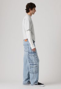 Ανοιχτό γκρι κοντομάνικο t-shirt φορεμένο πάνω από λευκό μακρυμάνικο πουκάμισο, συνδυασμένο με υπερμεγέθη ανοιχτό μπλε cargo τζιν με πολλές τσέπες. Μαύρα αθλητικά παπούτσια.