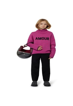 Pull en tricot rose avec le texte "AMOUR", pantalons noirs et chaussures noires texturées. Tenant un sac en forme de casque avec une finition sombre et brillante.