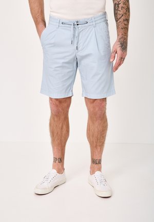 Man wearing light blue drawstring shorts, white sneakers, and white shirt, showing tattooed forearm and lower legs with text tattoos.