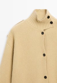 Beige ribbad cardigan med h�ög krage, som har svarta knappar och vida ärmar. Tyget har en texturerad, stickad look.