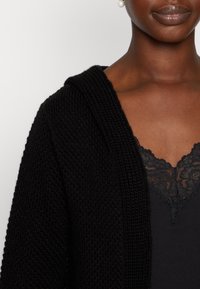 Cárdigan negro de punto con capucha, que presenta un acabado texturizado. Llevado sobre una blusa negra con borde de encaje, que revela intrincados detalles de encaje.