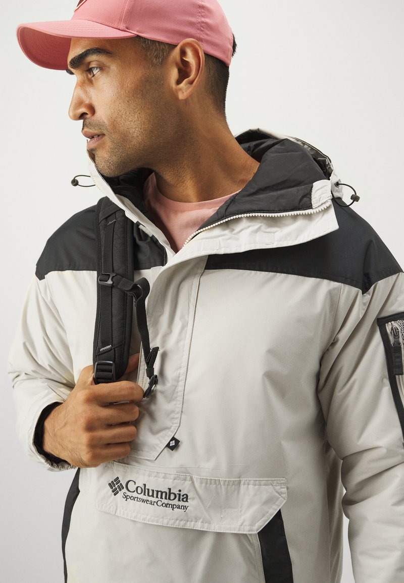 Lekki kurtka Columbia w białym kolorze z czarnymi akcentami, z przodem na pół zamka, regulowanym kapturem oraz teksturowaną kieszenią na ramieniu.