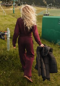 Bordový teplákový komplet s orezaným topom a nohavicami s širokými nohavicami; drží čiernu prešívanú bundu. Vo pozadí tráva a lanovka na zjazdovku.