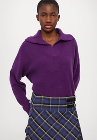 Pulover tricotat violet cu guler larg, asociat cu o fustă în carouri albastre, având accente galbene și roșii și un detaliu cu cataramă la talie.