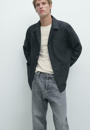 Giovane uomo con giacca nera abbottonata, maglietta bianca e jeans grigi sbiaditi, in piedi davanti a uno sfondo chiaro uniforme.