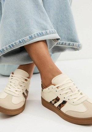 Pies con zapatillas beige con detalles de estampado de leopardo y suelas marrones, combinadas con vaqueros azul claro remangados.