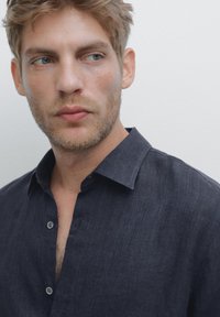 Blond man with light beard wearing an unbuttoned dark blue shirt, looking sideways against a plain light background.