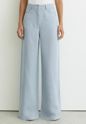 Femme portant un pantalon large taille haute bleu clair avec poches avant et fermeture par boutons, debout contre un mur blanc uni.