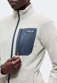 Κοντινό πλάνο ενός ατόμου που φοράει λευκό fleece μπουφάν με ζιγκ-ζαγκ σχέδιο, κλείνοντας μια ναυτικό μπλε τσέπη στο στήθος με το λογότυπο της Patagonia.