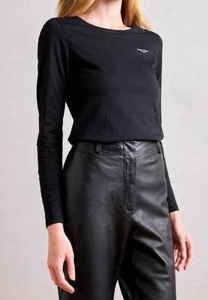 Kvinde iført sort, tætsiddende langærmet top med lille hvidt logo og sort, højtaljede læderbukser mod en ensfarvet baggrund.