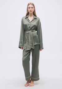 Γυναίκα που στέκεται ξυπόλητη φορώντας σετ πιτζάμες από σατέν σε ελιά πράσινο με λευκή ρίγα και μια ταιριαστή ζώνη δεμένη στη μέση.