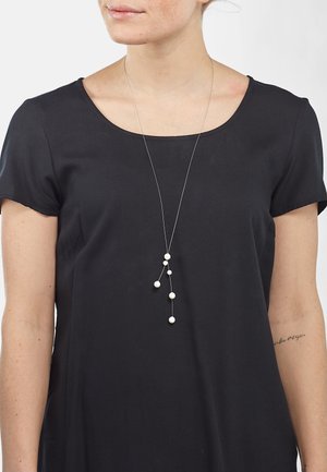 Silberfarbener Anhänger mit fünf unterschiedlich großen weißen Perlen, kaskadierende Gestaltung, getragen über einem schwarzen kurzärmeligen Oberteil. Einfach und elegant.