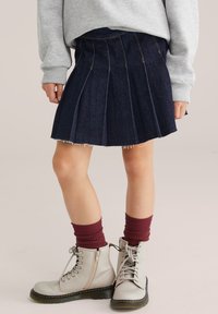 Denim plisirana krila v temno modri barvi z neobdelanim robom, v kombinaciji s svetlo sivimi čipkastimi čevlji in burgundskimi rebrastimi nogavicami. Teksturno bombažno blago.