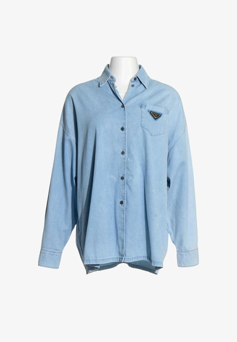 Chemise en denim bleu clair oversize à manches longues, boutonnée devant, col et un patch logo triangulaire sur la poche poitrine gauche.