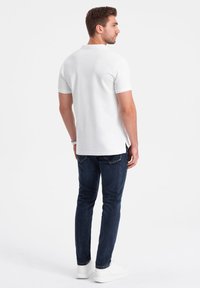 Biała koszulka polo z krótkim rękawem i klasycznym kołnierzykiem, w połączeniu z ciemnoniebieskimi dżinsami i białymi trampkami. Gładki materiał i luźny krój.