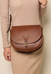 Borsa a tracolla in pelle marrone con forma arrotondata, dotata di chiusura a pattina, accessori in ottone e una tracolla intrecciata. Texture liscia con dettagli di cucitura.