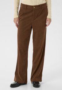Pantalons larges en velours côtelé marron avec une finition texturée, fermeture à boutons et poches. Associés à des chaussures noires pour un contraste de style.