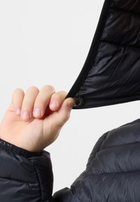 Sort quiltet jakke med blødt foer i hætten, med trykknaplukning og elastiske manchetter. Hånd holder om hættens kant, der afslører den indvendige tekstur.